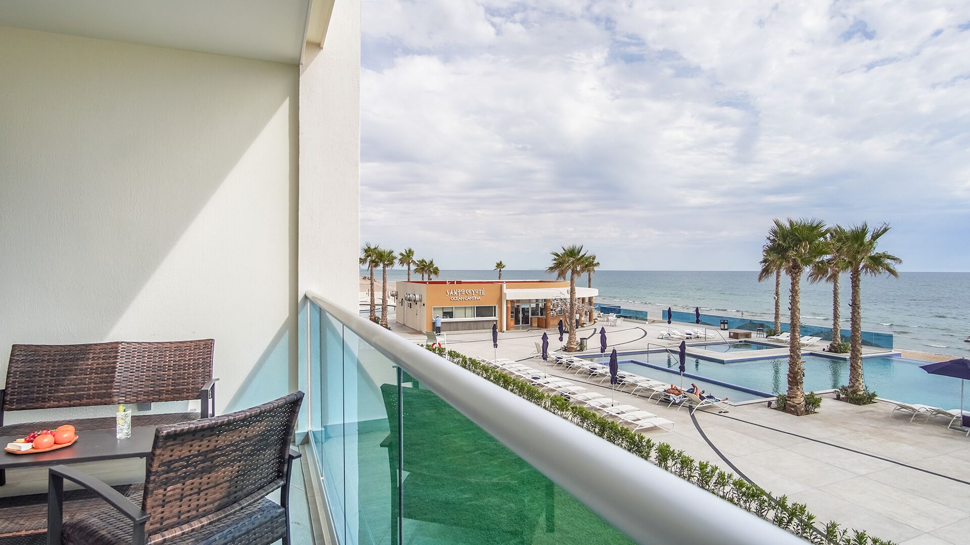 A balcony with wicker furniture, a pool area with lounge chairs, and a view of the beach and ocean.