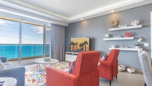 Una sala de estar moderna con un gran ventanal con vista al mar, un televisor de pantalla plana, sillas rojas y estanterías decorativas en la pared.