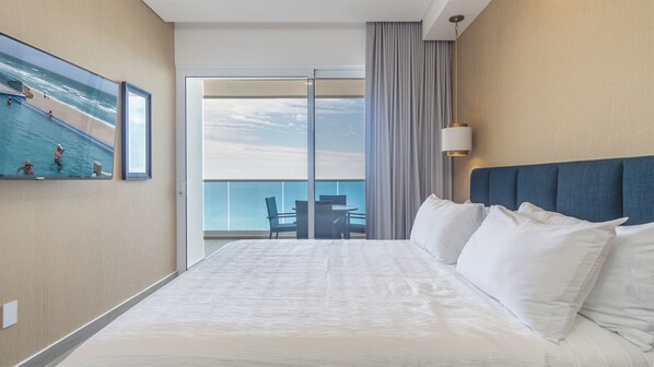 A hotel room with a large bed, a view of the ocean, a balcony with a table and chairs, and a painting of a beach scene.