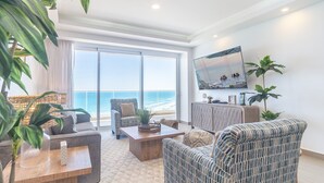 Ein großzügiges Wohnzimmer mit einer großen Schiebeglaswand, die auf einen Balkon mit Meerblick führt. Es gibt eine gemütliche Sitzecke mit einer Couch und Sesseln, einem Couchtisch und einem an der Wand befestigten Fernseher.