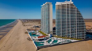 A beachfront hotel with two high-rise buildings, a swimming pool, and a desert landscape in the background.
