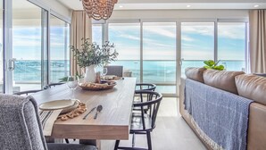 A modern dining area with a wooden table, chairs, and a view of the sea through large windows.
