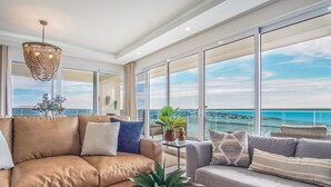 A spacious living room with a large window offering a view of the ocean, a brown leather sofa, a grey sofa, and a hanging light fixture.