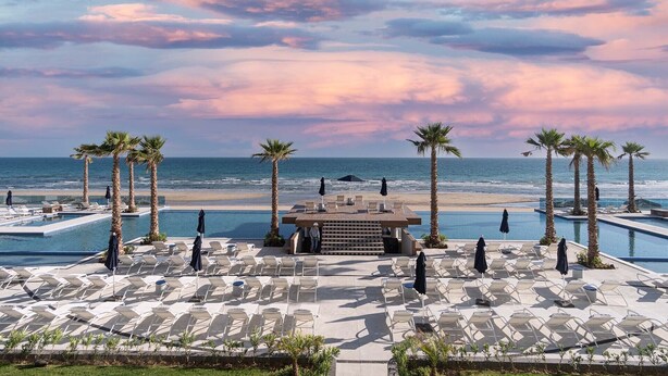 A resort pool area with lounge chairs and umbrellas, palm trees, and a view of the ocean.