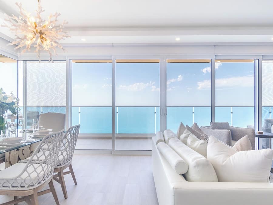 A modern living room with a large window overlooking the sea, a dining area with a glass table, and a white sofa with multiple cushions.