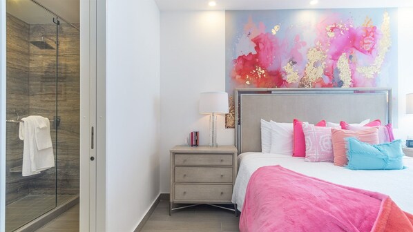 A modern bedroom with a glass shower, a bedside table with books, and a colorful abstract painting above the bed.