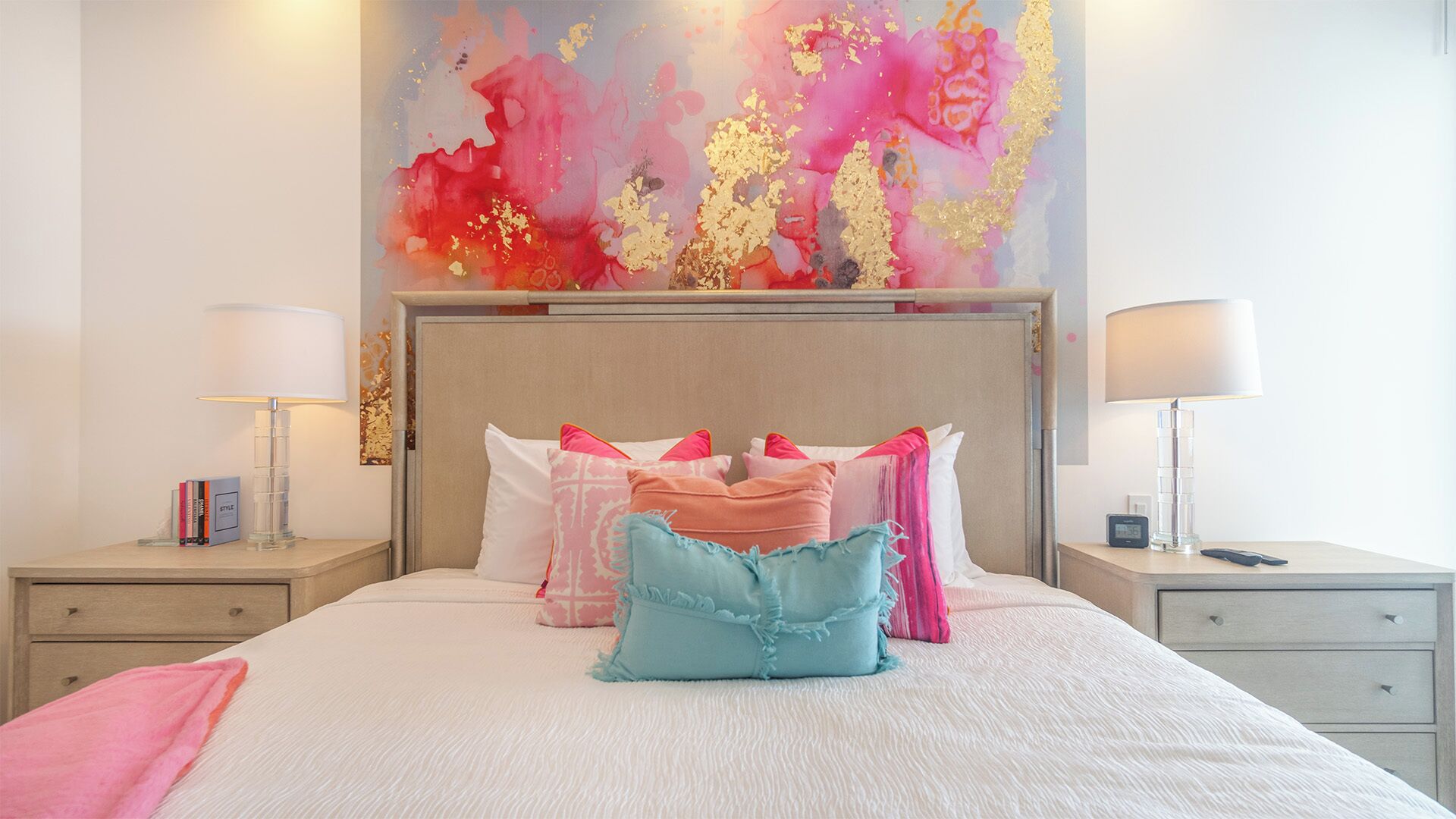 Una cama bien hecha con varias almohadas, una mesita de noche con una lámpara y una pared decorada con un cuadro abstracto colorido.