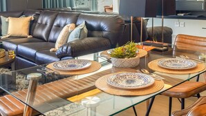 Un salon moderne avec un canapé en cuir, une table à manger en verre mise avec de l’argenterie et un centre de table, et un téléviseur en arrière-plan.