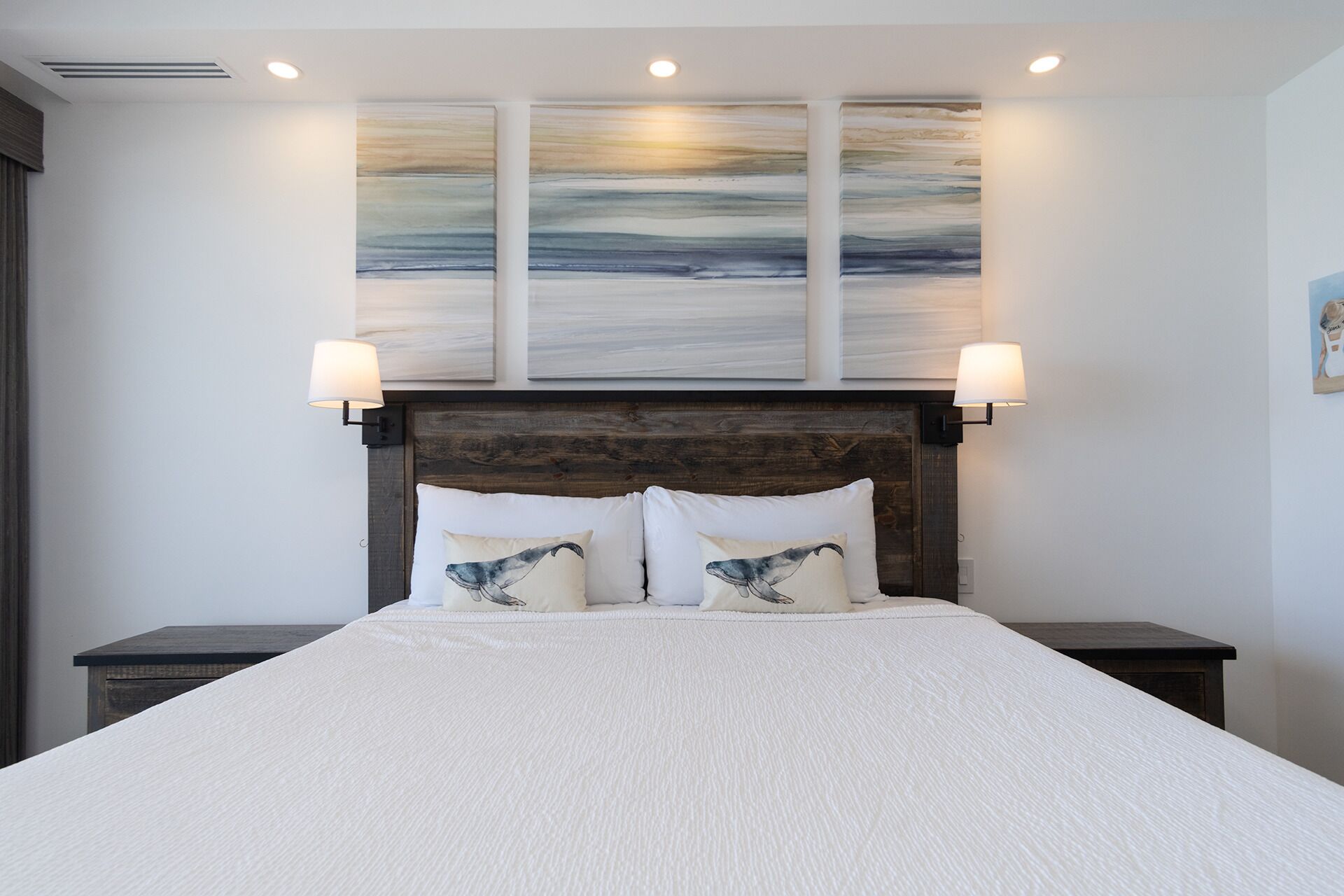 A neatly made bed with a white bedspread and two pillows with whale prints, flanked by two bedside tables with lamps, against a wall with three abstract paintings.