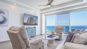 A spacious living room with a high ceiling, a ceiling fan, a large window with a view of the ocean, and a flat-screen TV mounted on the wall. - Perfect Family 2 Bedroom Condo E1804 Encantame (Puerto Peñasco)