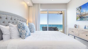 A bedroom with a large bed, a view of the ocean, a ceiling fan, and a TV on the wall.