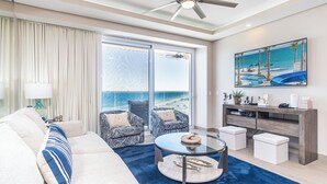 A modern living room with a sofa, two patterned armchairs, a glass coffee table, and a TV on a wooden console. A large window offers a view of the beach and ocean.
