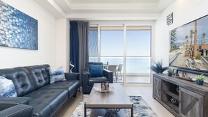 A modern living room with a leather sofa, a coffee table, and a view of the ocean through large windows. - Encantame Towers Viento 1103 (Puerto Peñasco)