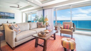 Ein modernes Wohnzimmer mit Sofa, Sessel und Hocker, einem Couchtisch und Blick auf den Ozean durch große Fenster.