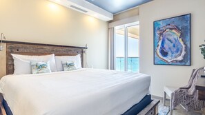A large bed with white linens, a wooden headboard, and decorative pillows. There is a large window with a view of the sky and a painting on the wall. - Panoramic Water Front Deluxe Condo Encantame Viento 605 (Puerto Peñasco)