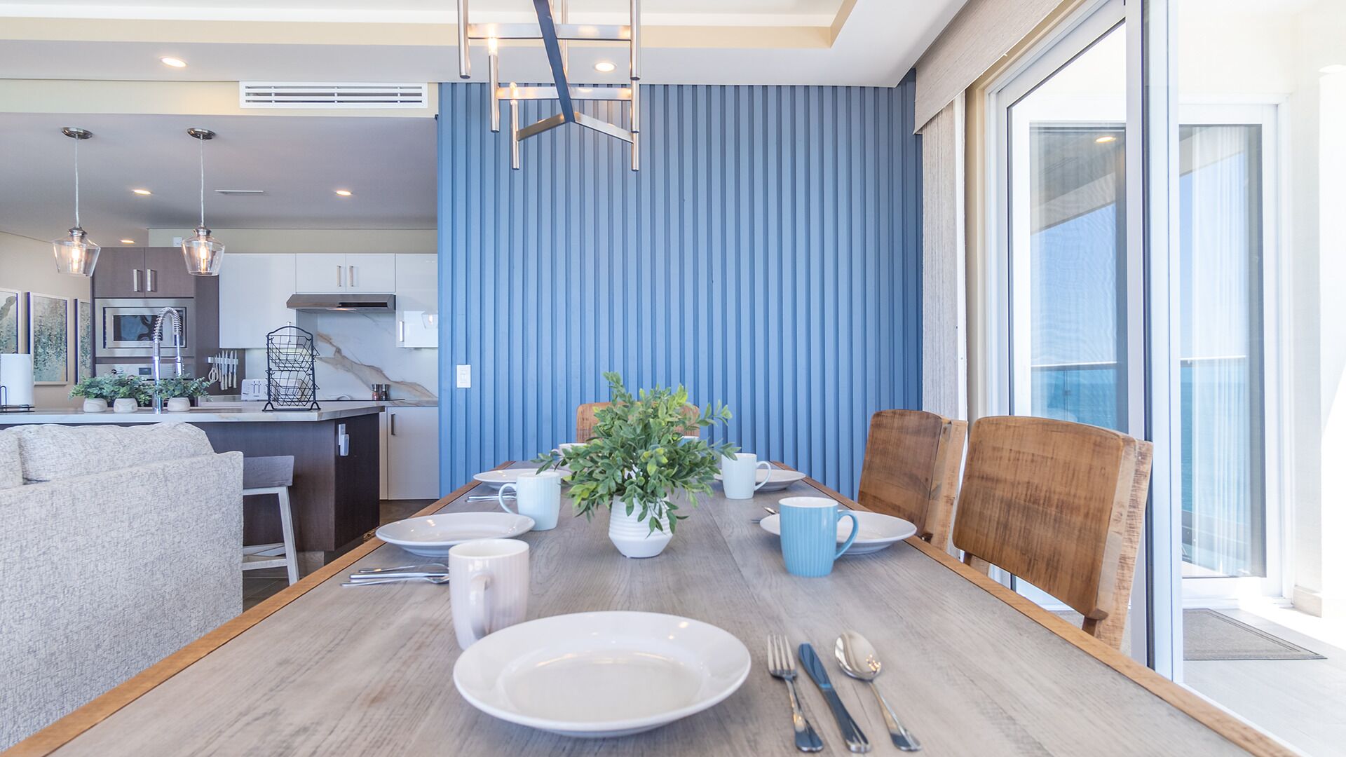 Un espace de repas moderne avec une table en bois dressée pour quatre personnes, un mur bleu et une vue sur la ville grâce à de grandes fenêtres.