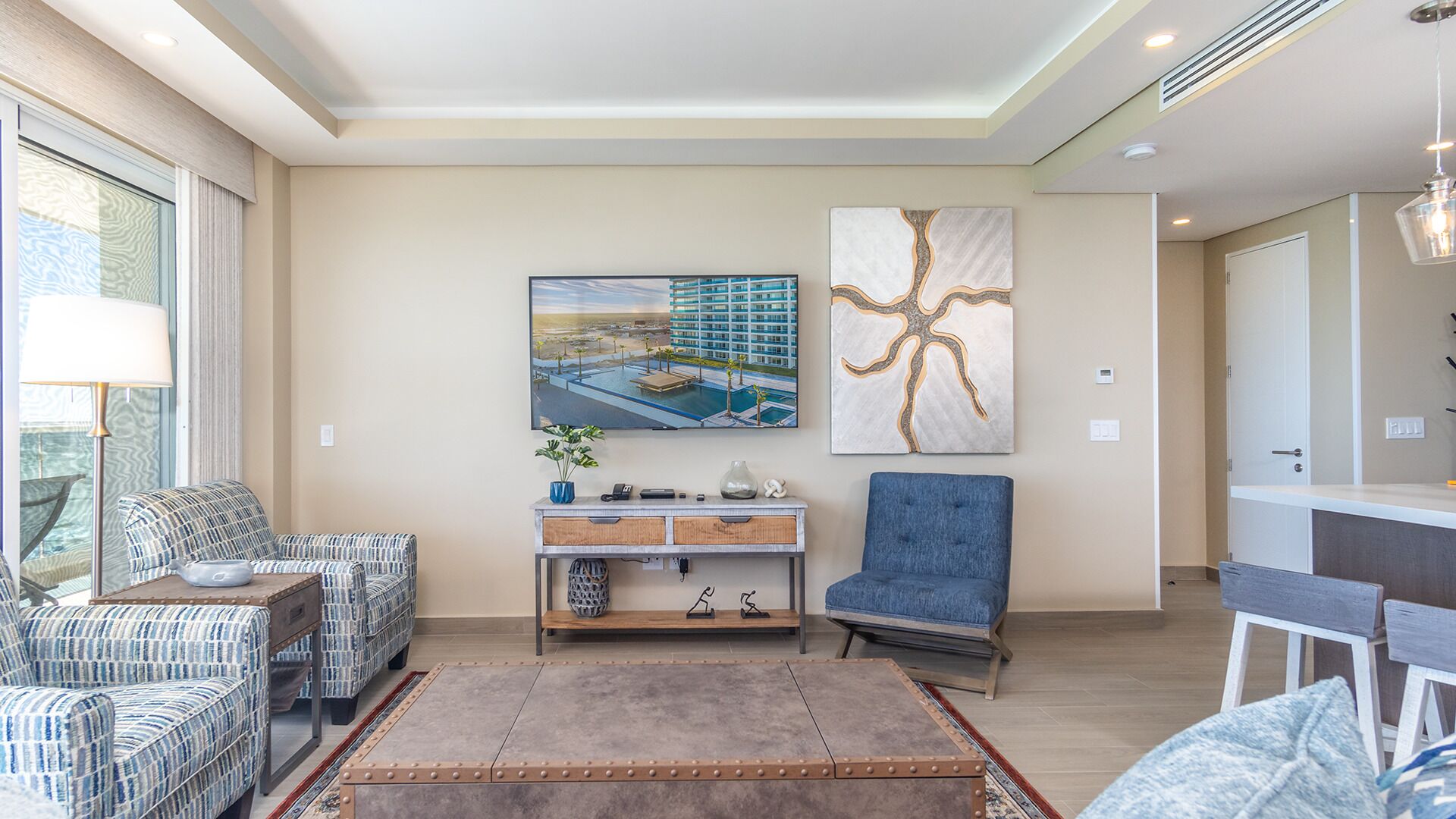 A modern living room with a sofa, armchair, and a coffee table. There is a painting of a building and a tree on the wall.