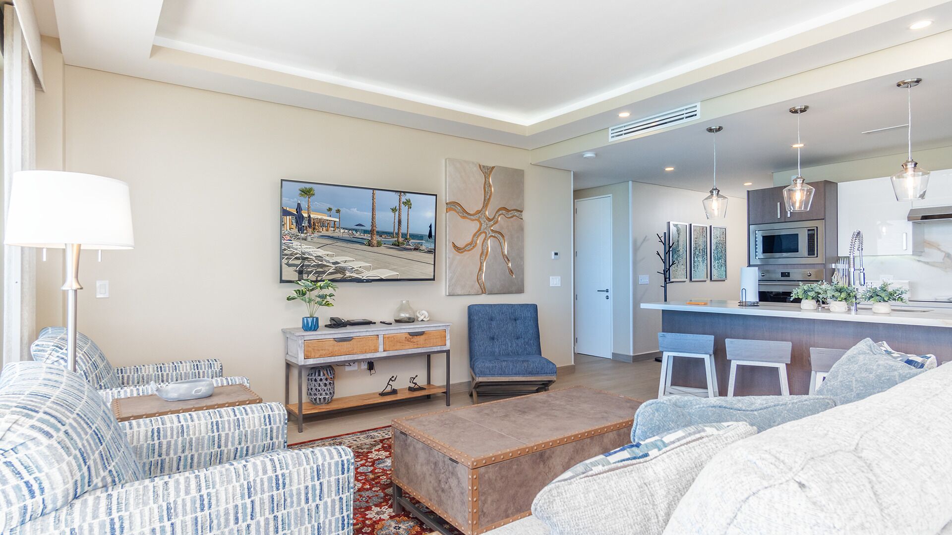 A modern living room with a sofa, a coffee table, and a TV. There is a kitchen area with a microwave and a sink.