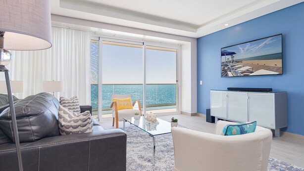 A living room with a sofa, a coffee table, a TV stand, and a painting of a beach scene. - Splash in The New Lazy River and Sleep In Luxury Encantame C-1104 (Puerto Peñasco)