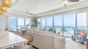 A spacious living area with a large window overlooking the ocean, a ceiling fan, and a dining area with wicker chairs. - Unique Condo by the Beach at Encantame Towers C305 (Puerto Peñasco)