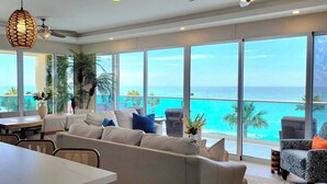 A modern living room with a large window overlooking a beach. There is a sofa, a dining table, and wicker chairs. - Unique Condo by the Beach at Encantame Towers C305 (Puerto Peñasco)
