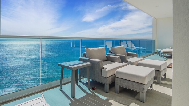 Een balkon met glazen hekwerk met uitzicht op een wateroppervlak. Er staan verschillende stoelen en een tafeltje.