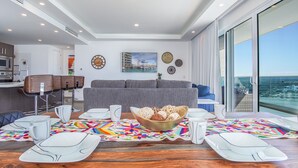 A modern living room with a grey sofa, a dining table set with white plates and cups, and a large window offering a view of the sea. - Beautiful 2 Bedroom Condominium Fully Equipped at the New Encantame Towers Verano 805 (Puerto Peñasco)