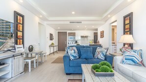 Un salon moderne avec un canapé bleu, des fauteuils blancs et une table basse agrémentée d’un élément décoratif vert. On aperçoit une cuisine au fond.