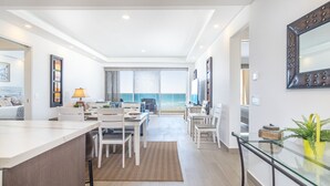 Un salon moderne doté d’une grande fenêtre donnant sur la plage, un coin repas avec des meubles blancs et une cuisine avec un îlot central en verre.