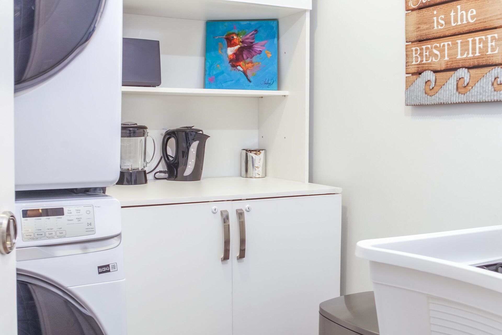 A laundry area with a washing machine, a dryer, a kettle, and a shelf with a painting and a sign.