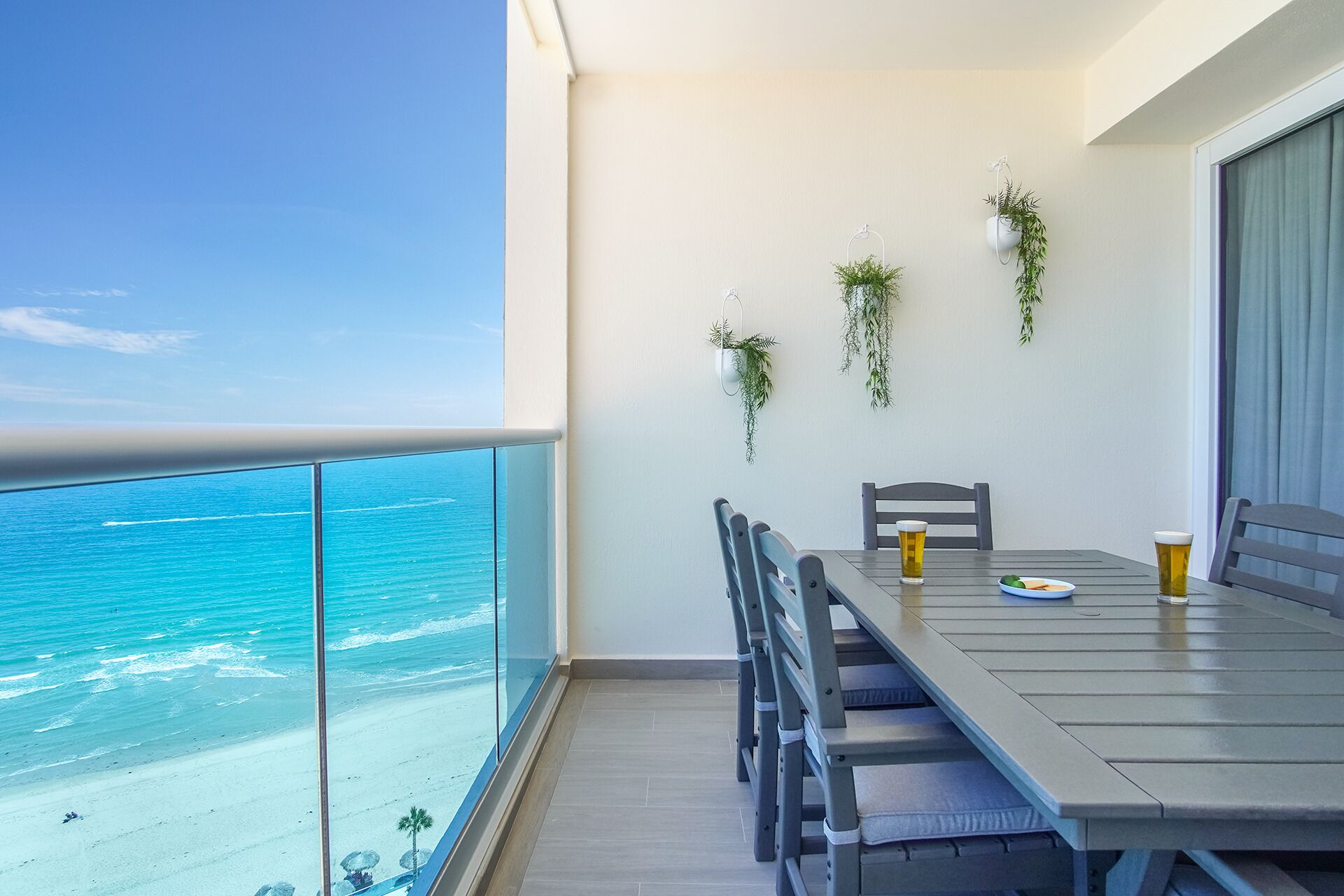 A balcony with a view of the beach and ocean. There is a table with chairs and two glasses of beer.