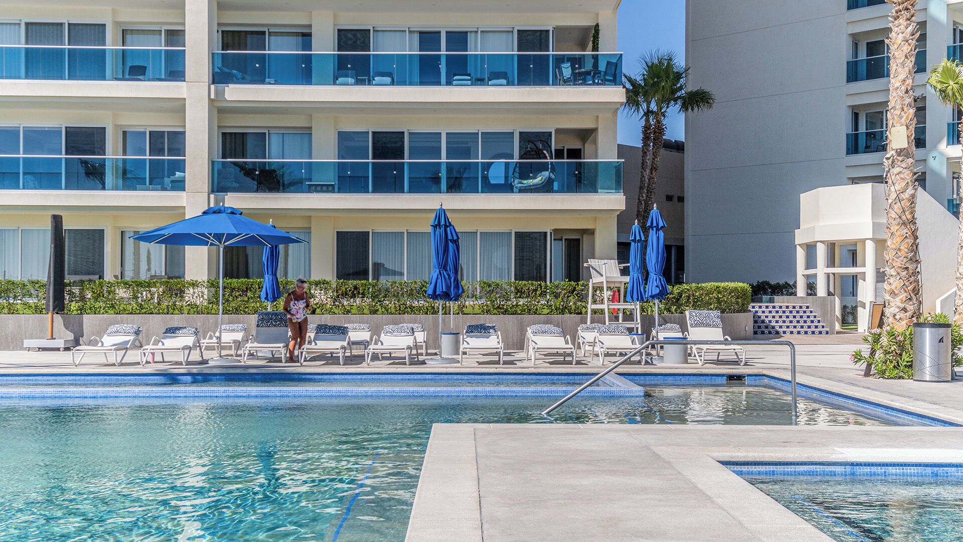 A hotel pool area with lounge chairs and umbrellas, a clear pool, and a building with multiple balconies.