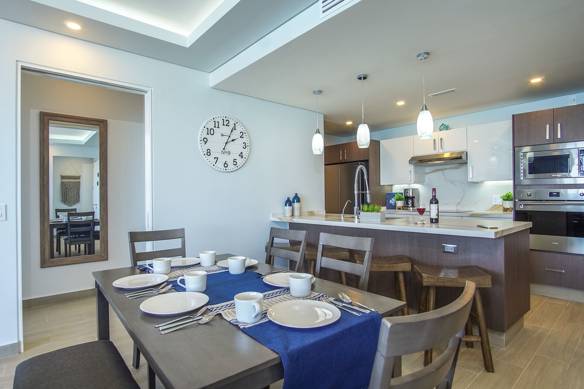 Une cuisine moderne avec un îlot central, un coin repas avec une nappe bleue et une horloge murale.