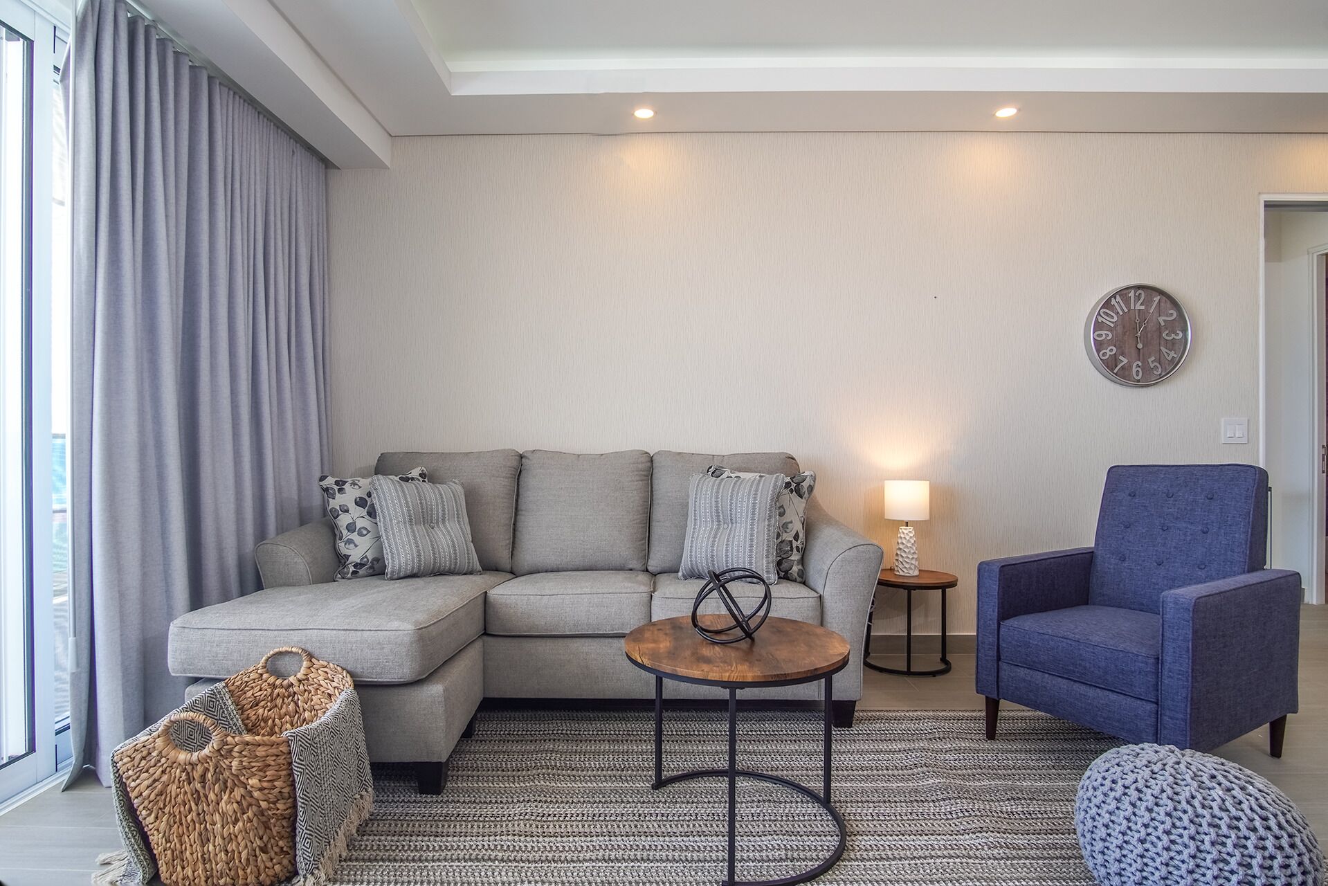 Ein modernes Wohnzimmer mit einem grauen Sofa, einem runden Holztisch, einem blauen Sessel und einem gewebten Pouf.