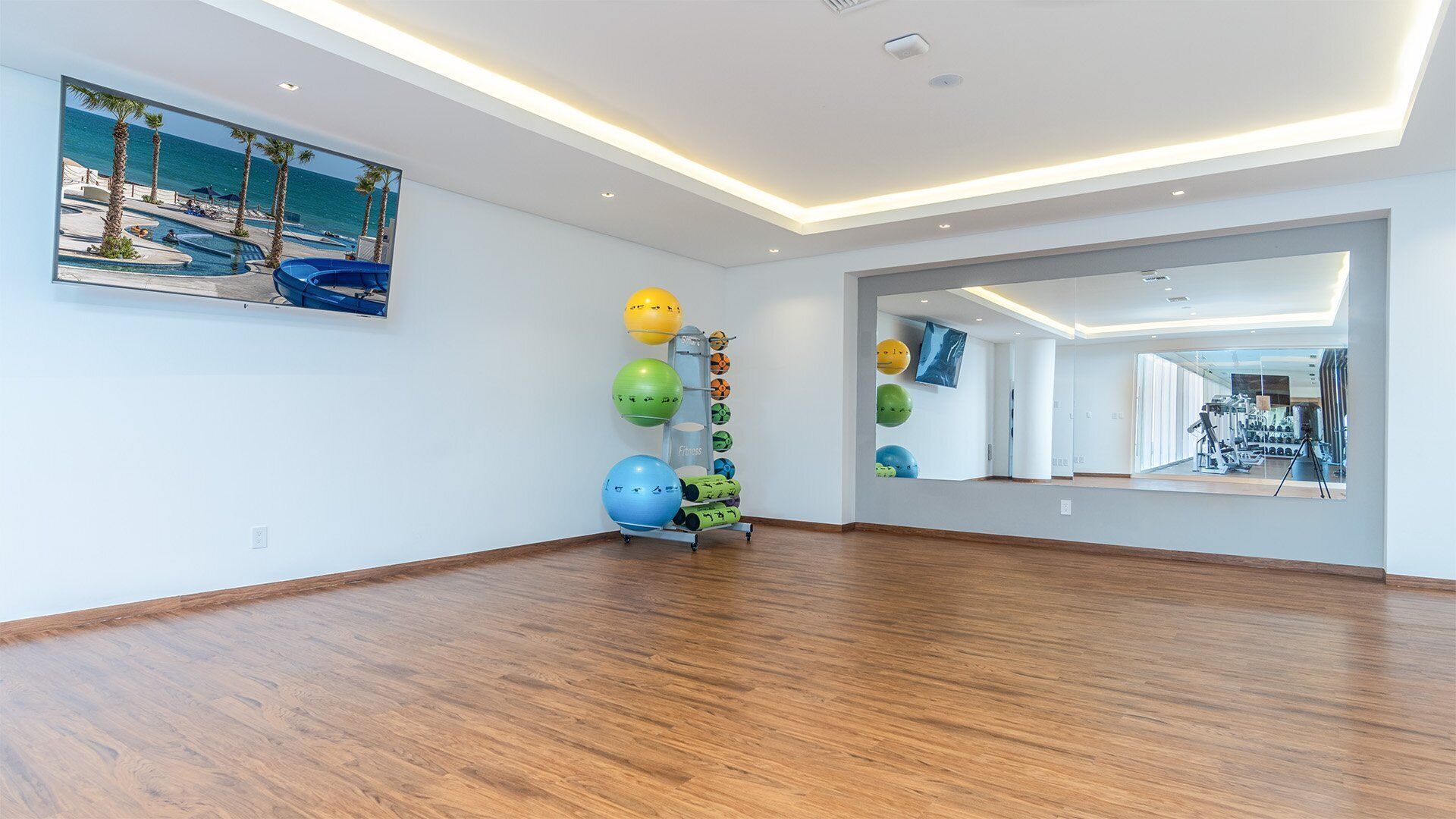 Um quarto espaçoso com piso de madeira, um espelho grande refletindo uma área de ginástica e uma pintura com uma cena de praia na parede.