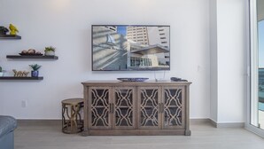 A modern living room with a large window, a flat-screen TV mounted on the wall, a wooden cabinet with glass doors, and decorative items on shelves. - Looking for the Perfect Scape From the City? Peaceful 2 Bedroom Condo Verano 804 (Puerto Peñasco)