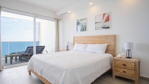 A bedroom with a large bed, a wooden nightstand, a chair, and a view of the sea through a sliding door. - Stunning 2BR Condo at Encantame Towers Velero 2302 in Playa Encanto (Puerto Peñasco)