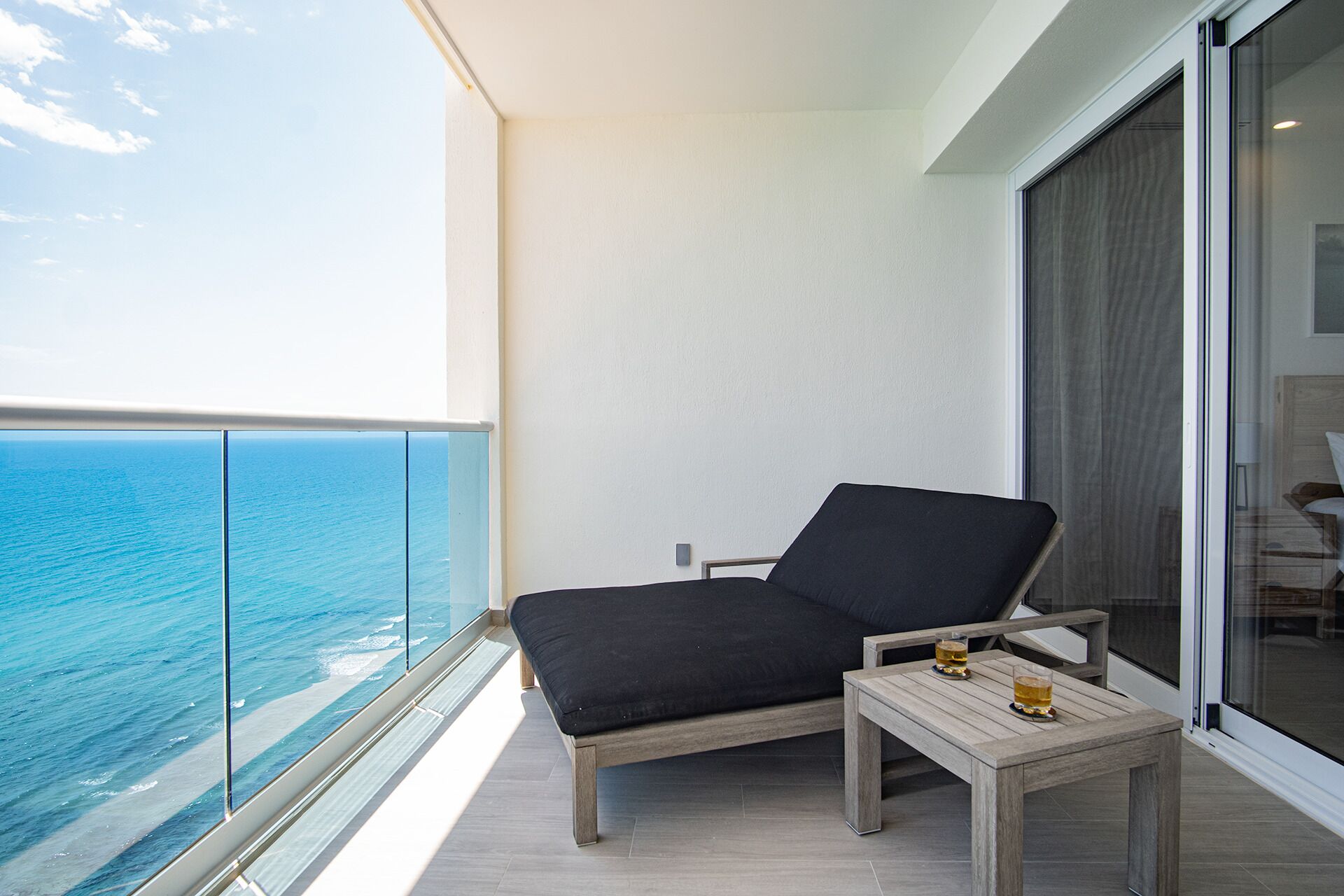 A balcony with a view of the ocean, a black lounge chair, and a small wooden table with two glasses.