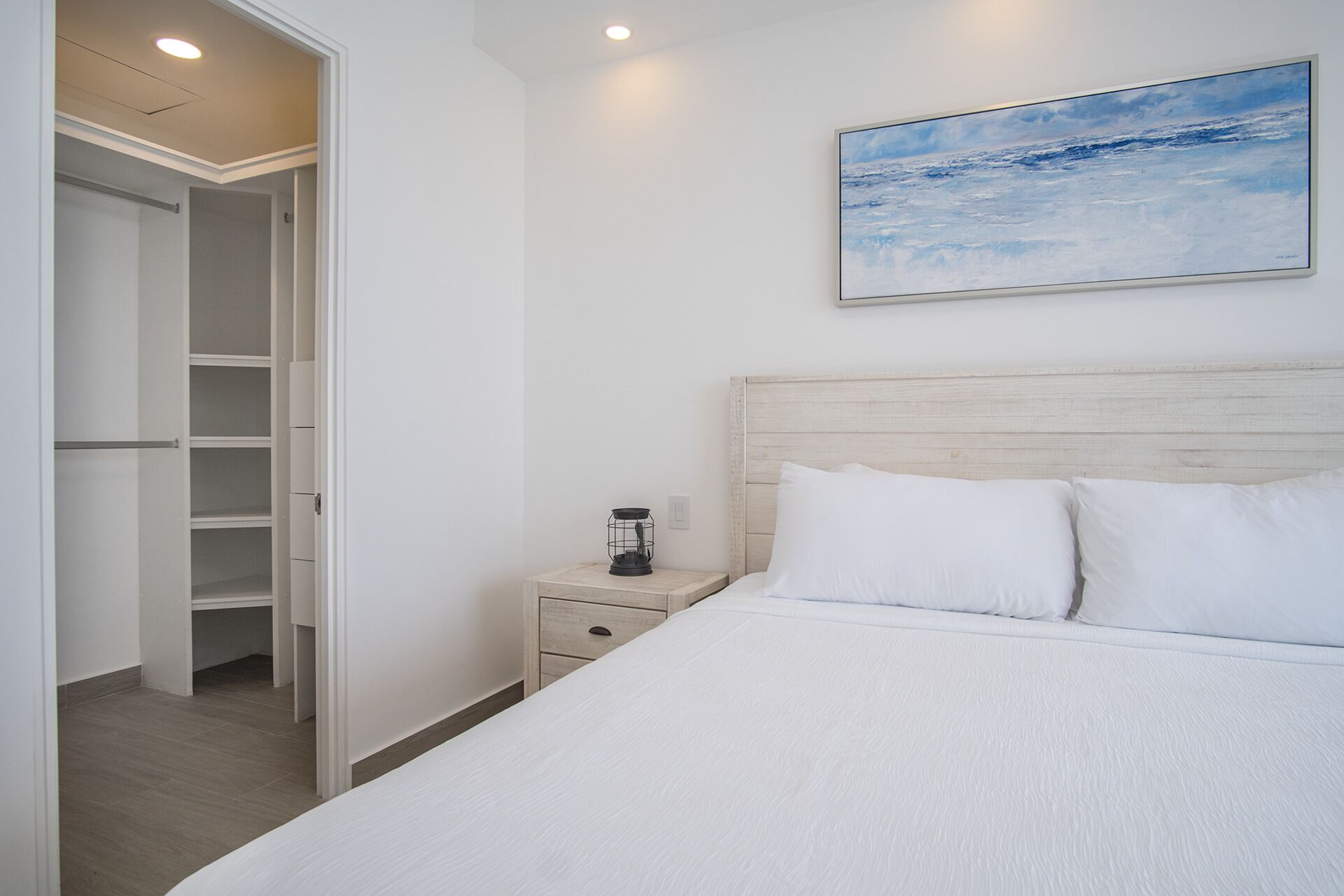A bedroom with a bed, a nightstand, a lamp, a painting, and a walk-in closet.