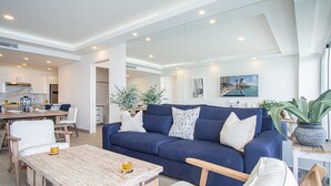 A modern living room with a blue sofa, white cushions, and a wooden coffee table. There is a dining area with a wooden table and chairs, and a kitchen in the background. - Stunning 2BR Condo at Encantame Towers Velero 2302 in Playa Encanto (Puerto Peñasco)