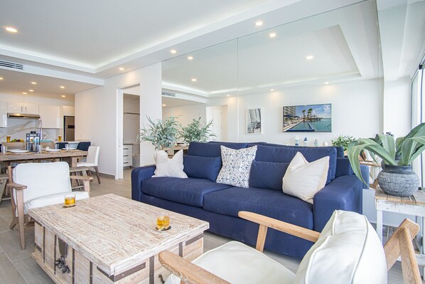 Una sala moderna con un sofá azul, cojines blancos y una mesa de centro de madera. Se observa un comedor con una mesa y sillas de madera, y una cocina al fondo.