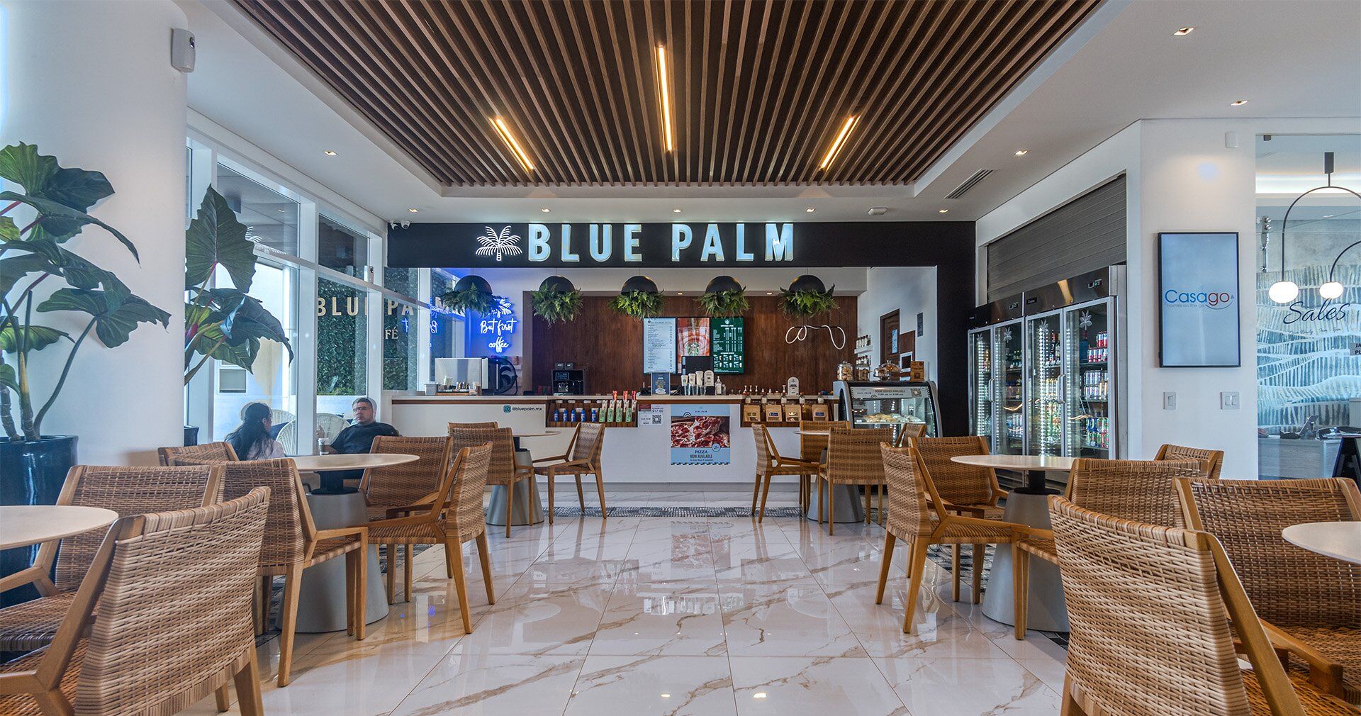 A modern bar area with a sign reading "BLUE PALM" and a counter with various items. There are wicker chairs and tables, and a large window with plants.