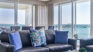 A modern living room with a leather sofa, blue cushions, and a glass coffee table. Large windows offer a view of the sea and a distant cityscape. - Awesome 3 Bedroom Condo Encantame Tower E 201 (Puerto Peñasco)