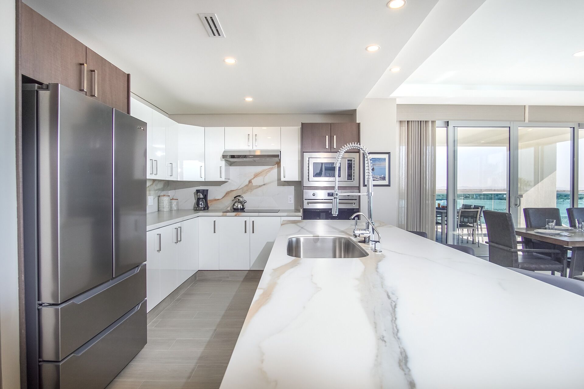 A modern kitchen with stainless steel appliances, a marble countertop, and a view of the ocean through large sliding doors.