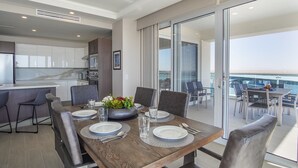 A dining area with a wooden table set for four, a kitchen in the background, and a view of the sea through large glass doors. - Awesome 3 Bedroom Condo Encantame Tower E 201 (Puerto Peñasco)
