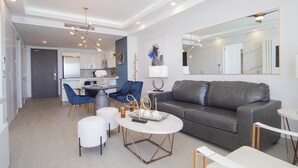 A modern living room with a grey sofa, two blue chairs, a white coffee table, and a dining area with a kitchen in the background. - Stay in Luxury/splash in New Lazy River This Winter Encanatame Towers 903 (Puerto Peñasco)