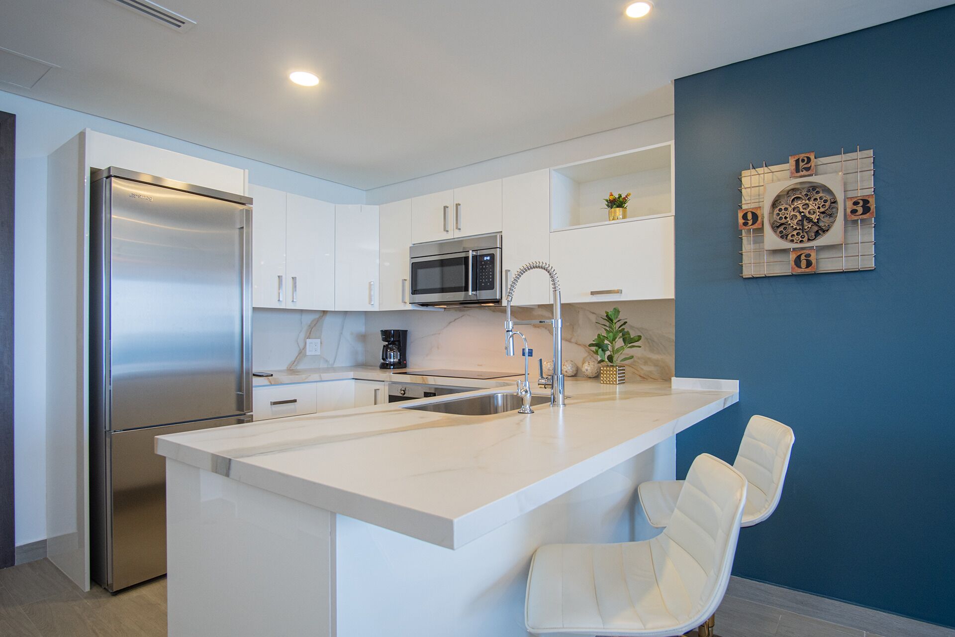 A modern kitchen with a stainless steel refrigerator, a microwave, and a sink with a flexible faucet. There is a white island with a chair and a decorative clock on the wall.