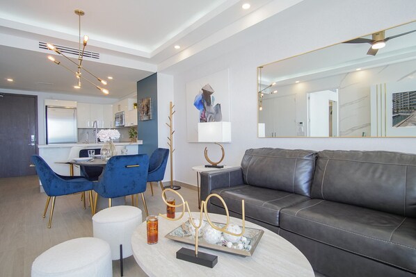 Una sala de estar moderna con un sofá de cuero, sillas azules, una mesa de centro y un comedor, con una cocina al fondo.