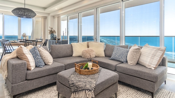 A modern living room with a gray sectional sofa, a coffee table with a candle and a basket, and a view of the ocean through large windows. - Wonderful 1 Bedroom Condominium Encantame Verano 2103 (Puerto Peñasco)