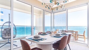 Un área de comedor con una mesa lista para cuatro personas, con vista al mar, con una silla colgante y vista del océano a través de grandes puertas de vidrio.